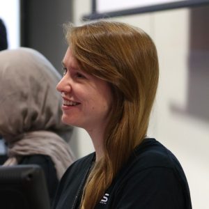 Person with long brown hair smiling, wearing a black shirt. Another person in a hijab is partially visible beside them.