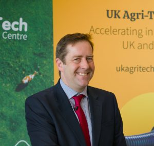 Smiling man in a suit with a maroon tie standing in front of a UK Agri-Tech banner.