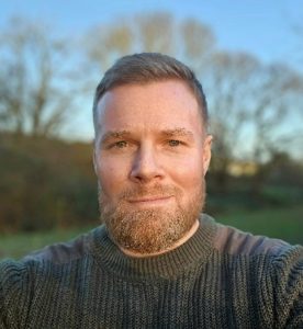 Man with a beard wearing a sweater, standing outdoors with trees in the background.