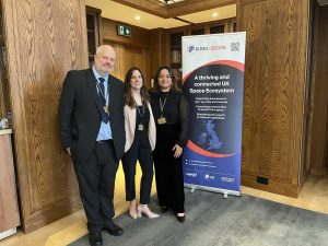 Three people standing and smiling beside a UK Space Ecosystem banner in a wood-paneled room.