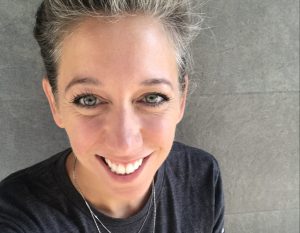 Smiling person with short hair wearing a dark shirt against a gray background.