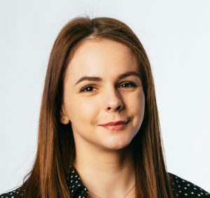 Smiling person with long brown hair, wearing a black polka-dot shirt, against a plain background.