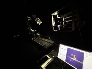A robotic arm in a dark room works with a small satellite. A laptop displays engineering software.