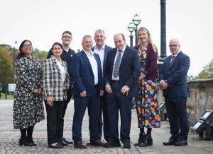 Eight people in business attire standing outside, smiling towards the camera, with lamp posts and trees in the background.
