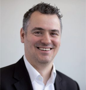 Smiling man with short gray hair in a white shirt and black suit jacket against a plain background.