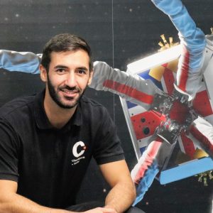 Smiling man in a black shirt sits in front of a space-themed mural featuring astronauts and a satellite.