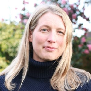 Woman with long blonde hair in a black turtleneck, standing in a garden with pink flowers in the background.