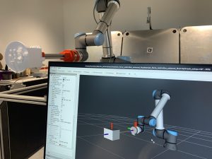 A robotic arm near a computer screen displaying a 3D model of the arm in a laboratory setting.