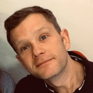 Man with short brown hair and light eyes, wearing a dark shirt, tilts his head while looking at the camera.