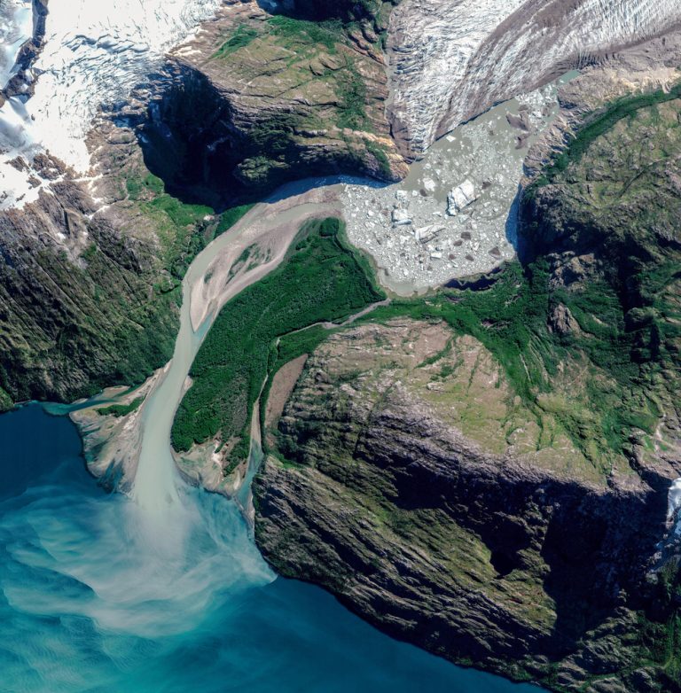 Aerial view of a glacier melt, creating a braided river flowing into a turquoise lake, surrounded by rocky terrain and greenery.