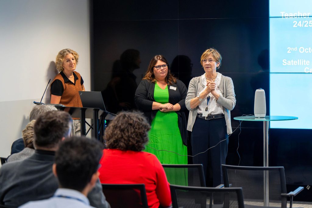 Three women present at a conference, addressing an audience in a room with a screen displaying information.