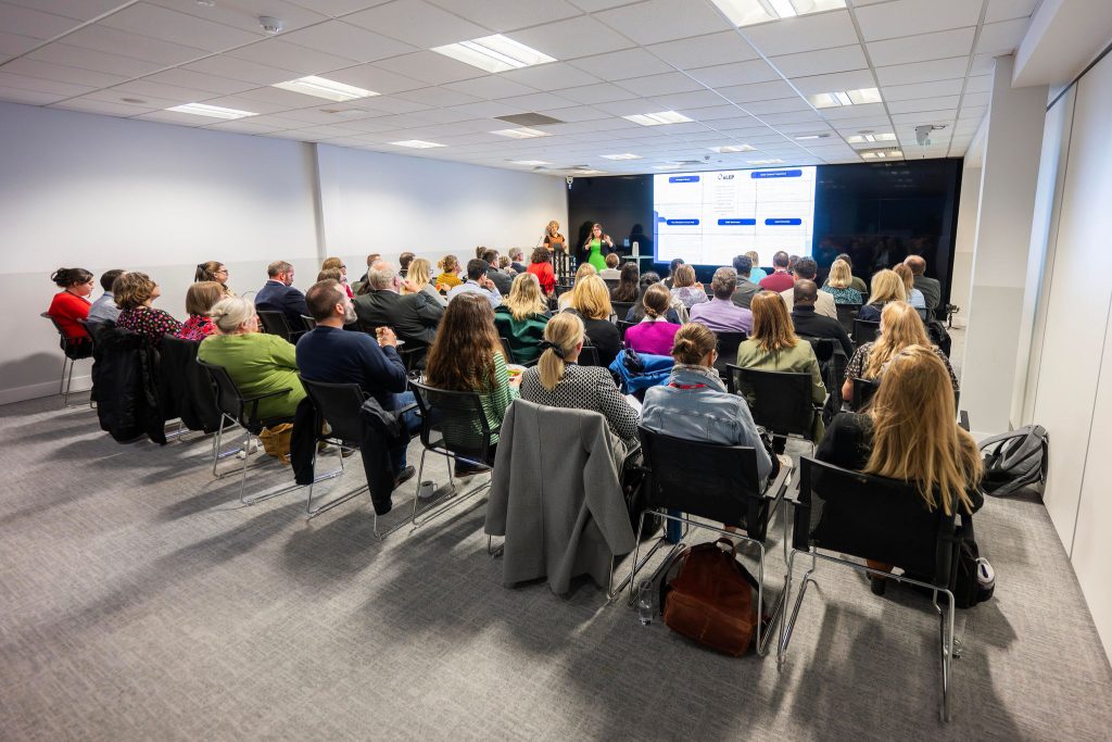A group of people seated in a conference room, listening to a presentation projected on a screen.