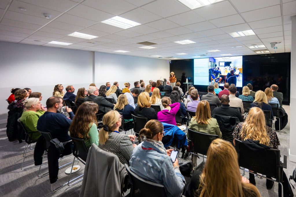 A group of people seated in a conference room watching a presentation on screens at the front.