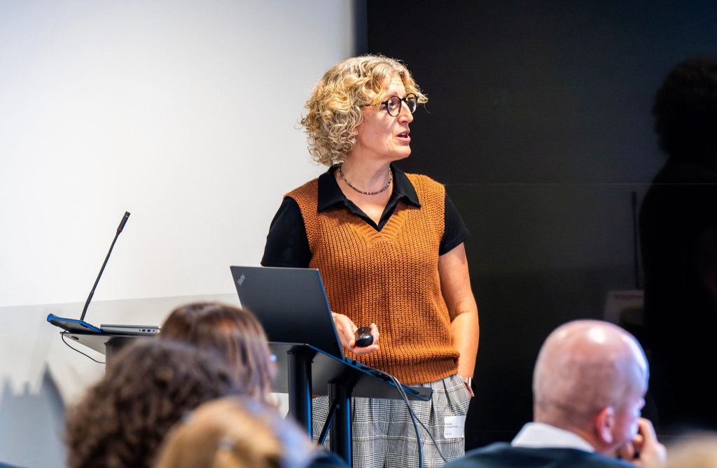 A person with curly hair speaks at a podium, using a laptop, in front of an audience.