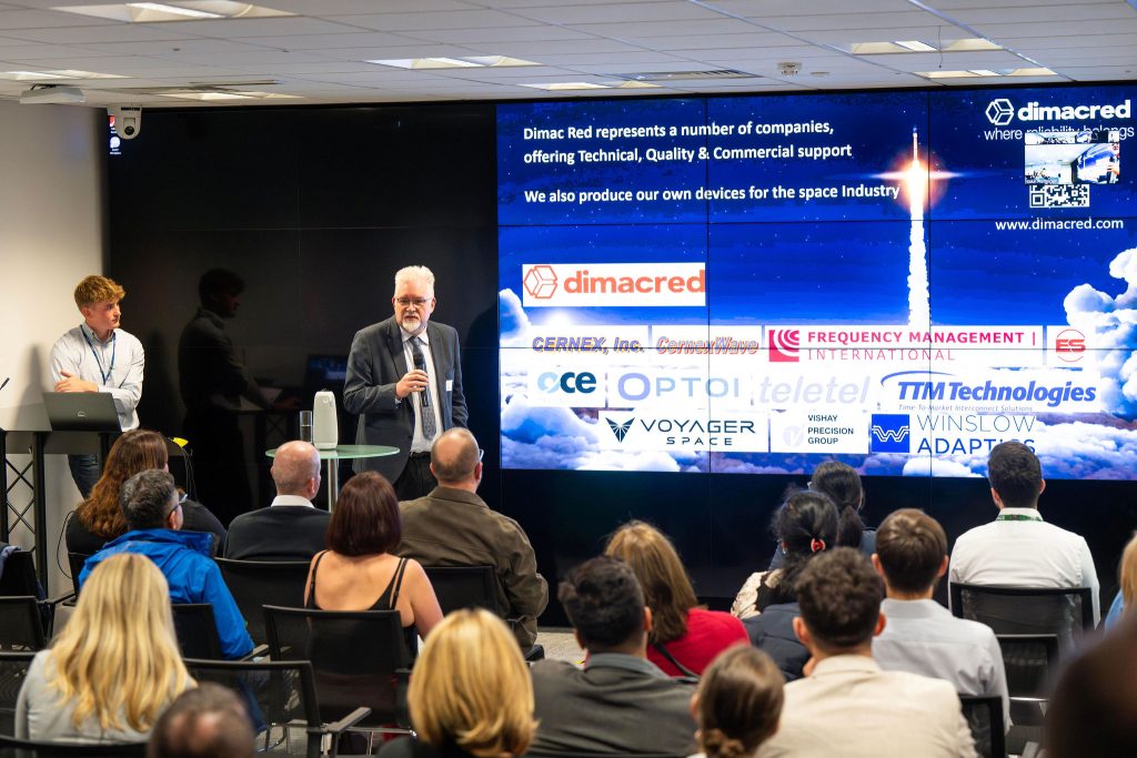 Speaker presenting to an audience with a display of various company logos related to the space industry behind him.