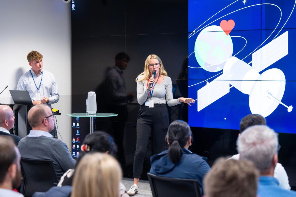 A woman presents near a large screen with satellite graphics, an audience and a man stand by.