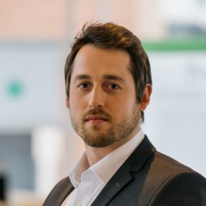 A man with short brown hair and a beard, wearing a suit jacket and white shirt, looks at the camera.