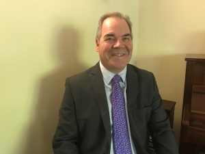 Smiling man in a suit and patterned tie sits indoors near a piano and a cream-colored wall.