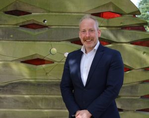 A man in a navy suit stands smiling in front of a modern outdoor sculpture and greenery.