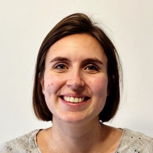 Smiling woman with straight brown hair and a light grey top against a plain white background.