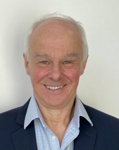 Smiling older man with short gray hair in a blue blazer and light blue collared shirt against a white background.