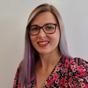 Smiling woman with glasses and long, straight hair, wearing a floral top, in front of a plain white background.