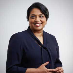 Woman with short black hair wearing a navy blouse, smiling and looking to the side against a light background.