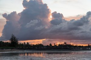 Large dramatic clouds over a beach at sunset, with people gathered near the shoreline.