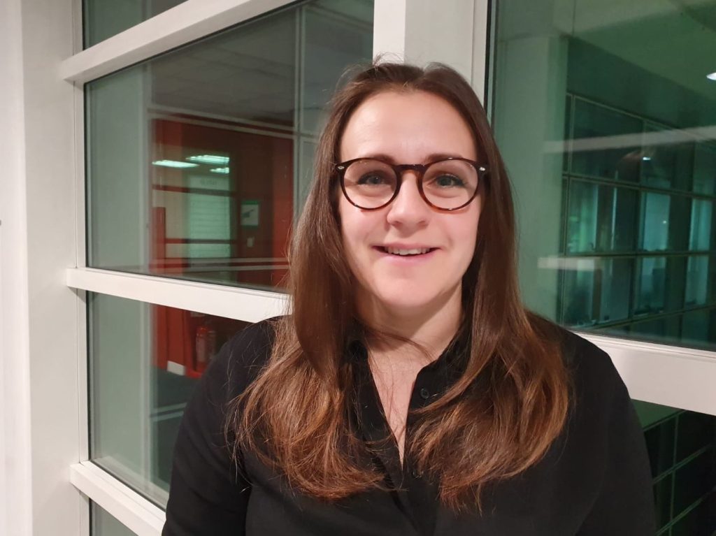 A woman with long brown hair and glasses smiles while standing indoors by large windows.