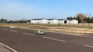 Two small remote-controlled airplanes on a runway with hangars and trees in the background.