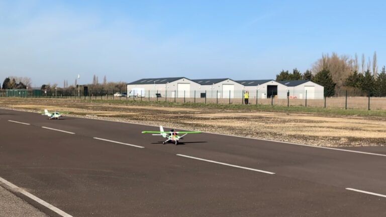 Two small remote-controlled airplanes on a runway with hangars and trees in the background.