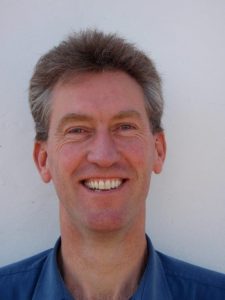 Smiling man with short, light brown hair wearing a blue collared shirt, standing in front of a white background.