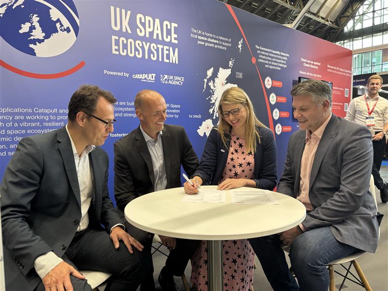 Four people sit at a round table signing documents at a UK Space Ecosystem event, smiling and engaged.