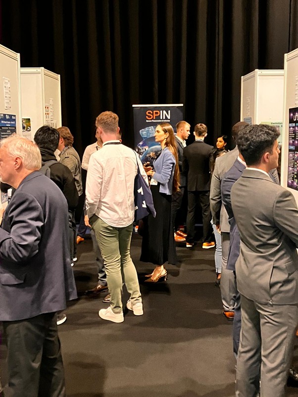 People in business attire interact and view posters at a busy conference or exhibition hall.