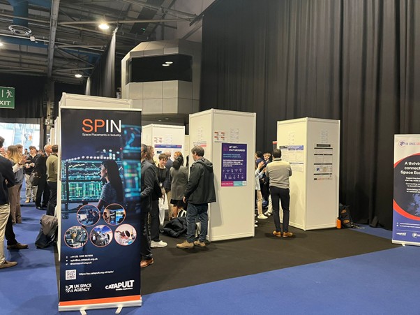 People gather at an exhibition with booths and banners, including one for SPIN Space Placements in Industry.