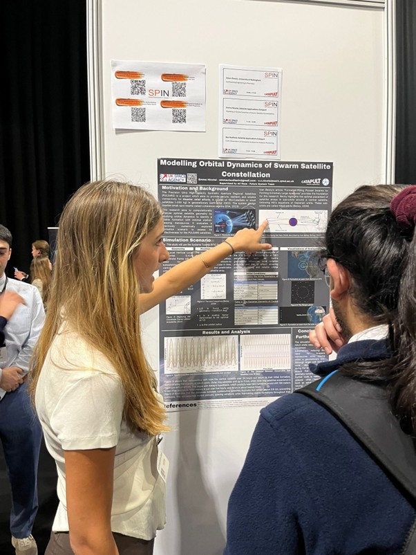 Woman presenting a scientific poster to another person at a conference, pointing at a chart on the poster.