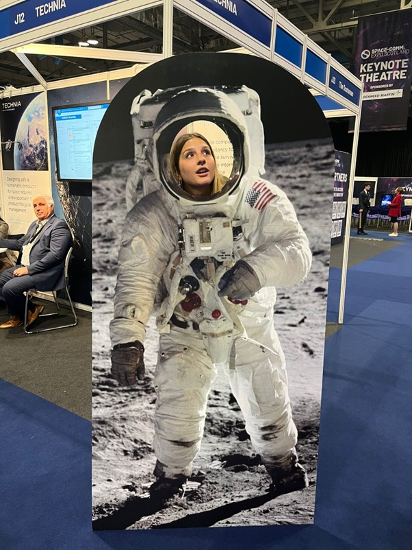 A woman poses with her face in a cardboard astronaut cutout at a tech expo booth.