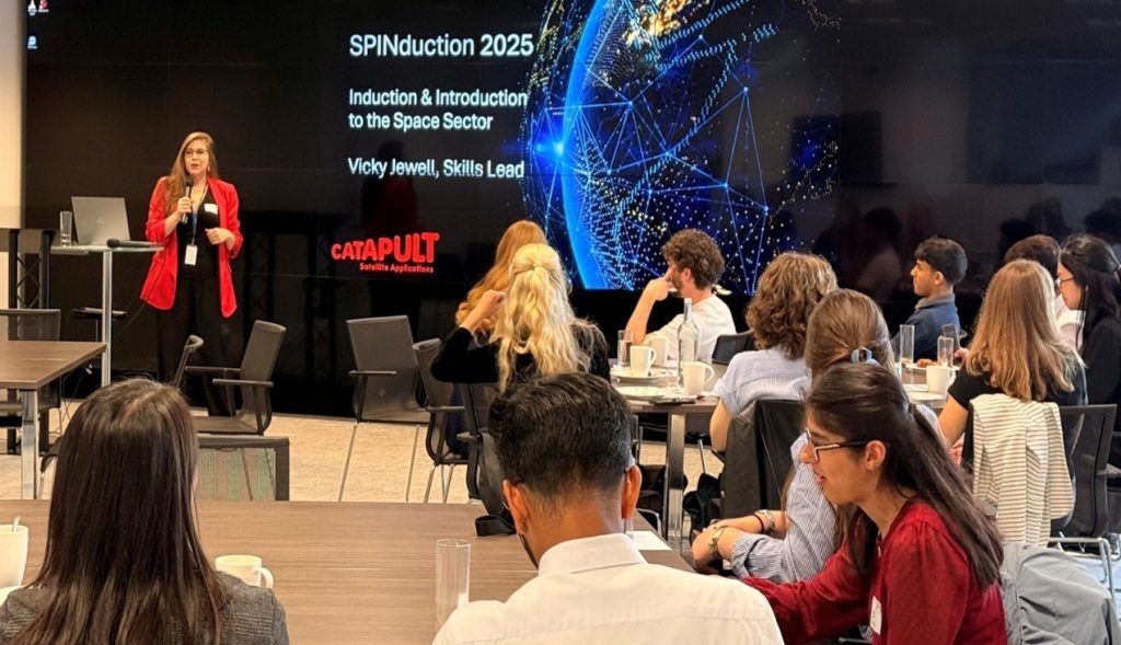 A woman presents "SPINduction 2025" to an audience in a conference room with a large screen behind her.