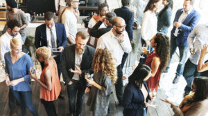 People at a social event mingling, talking, and holding drinks in a bright indoor setting.