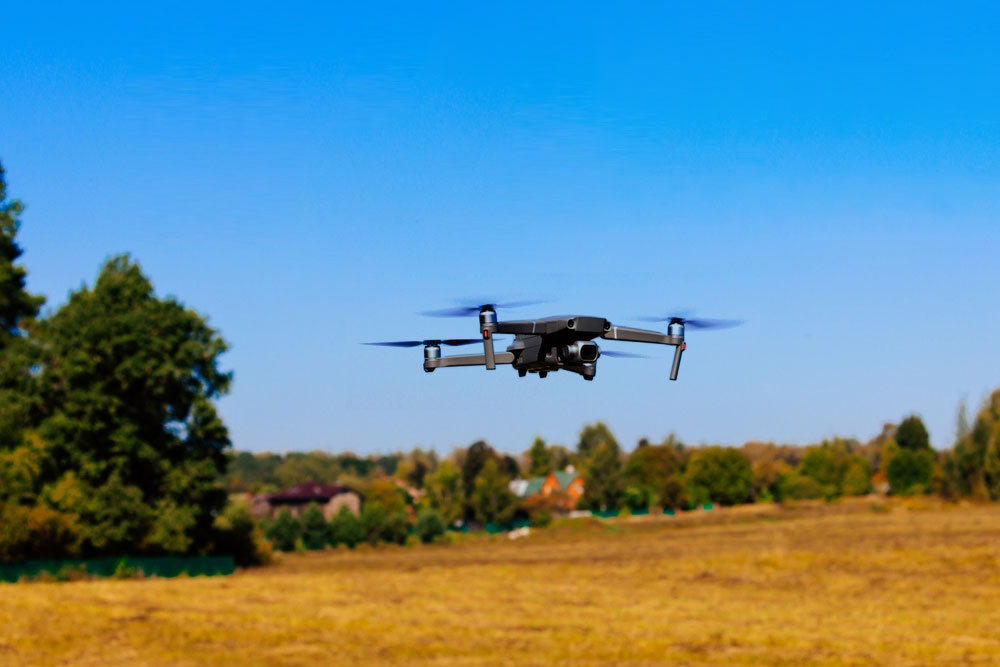 A black drone flying over an open field with trees and a clear blue sky in the background.