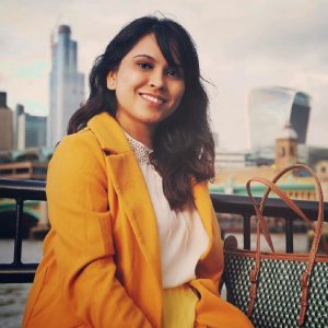 Woman in a yellow coat smiling outdoors with city buildings in the background.