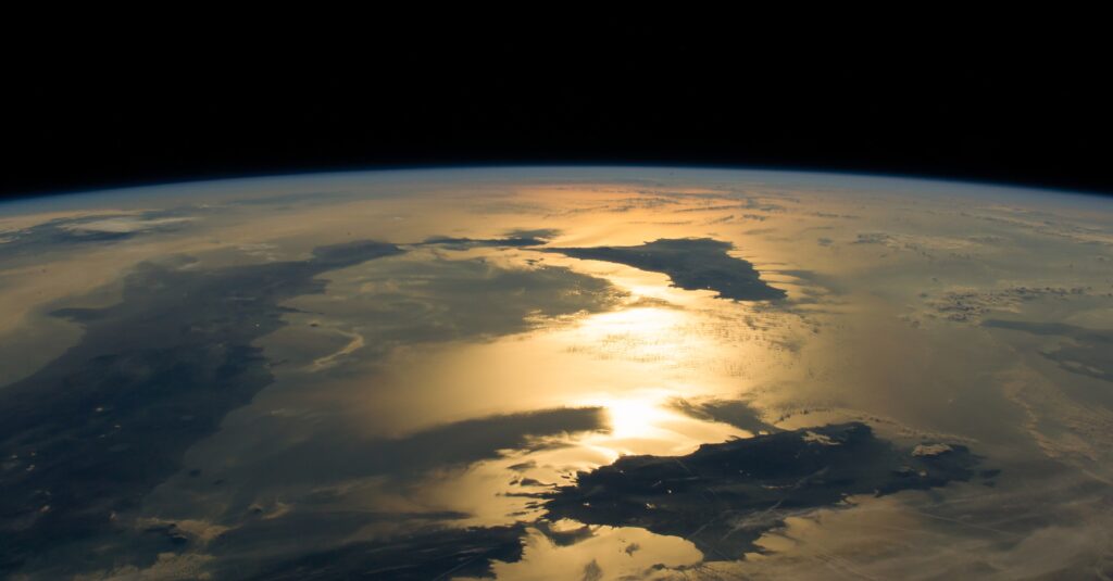 View of Earth’s surface with golden sunlight reflecting on water, seen from space with a dark horizon.