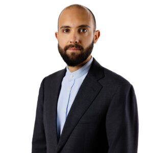 Man with a beard in a dark suit and light blue shirt, standing against a white background.