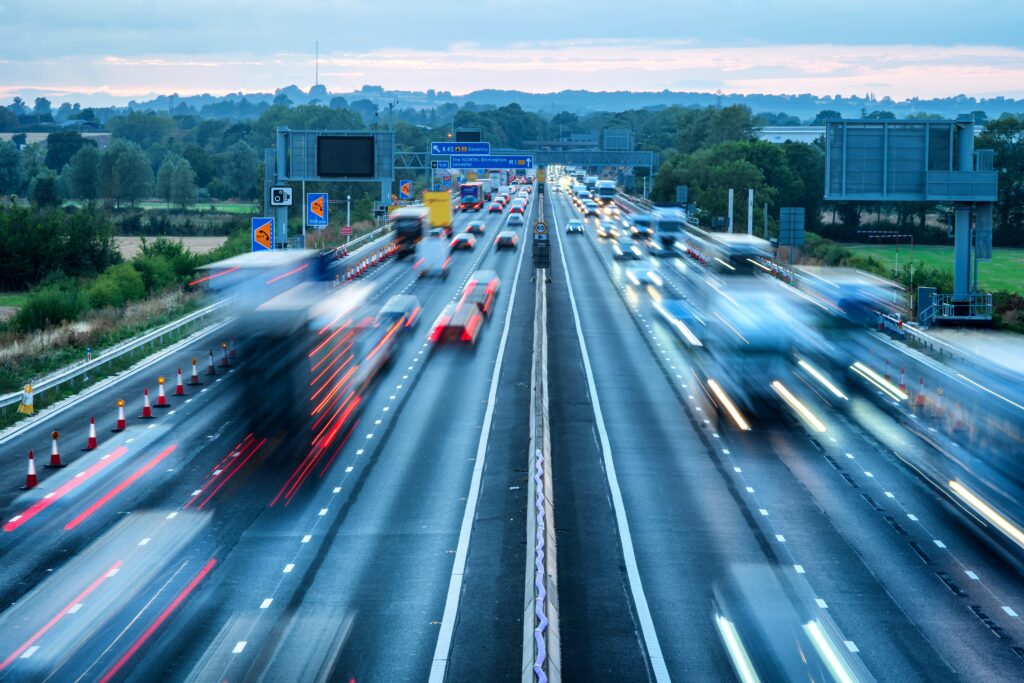 Blurred cars drive along a busy multi-lane highway surrounded by greenery at dusk.