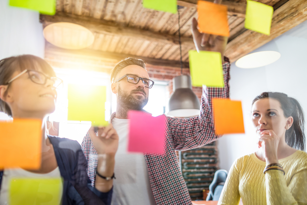 Three people brainstorming, placing colorful sticky notes on glass in a bright, modern office.