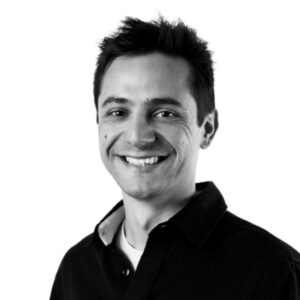 Black and white photo of a smiling man with short dark hair, wearing a collared shirt against a white background.