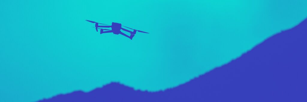 Silhouette of a drone flying against a bright blue sky with dark blue mountains below.