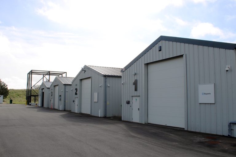 Row of grey industrial warehouses with large numbered doors on a paved lot, under a cloudy sky.