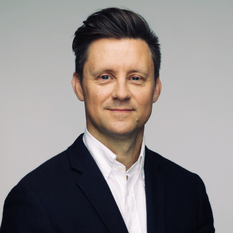 Man with short brown hair wearing a dark blazer and white shirt, smiling at the camera against a gray background.