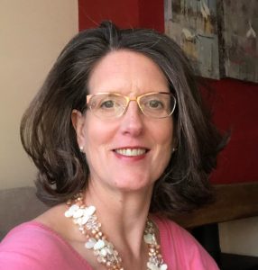 Woman with glasses and shoulder-length brown hair, wearing a pink top and floral necklace, smiling.